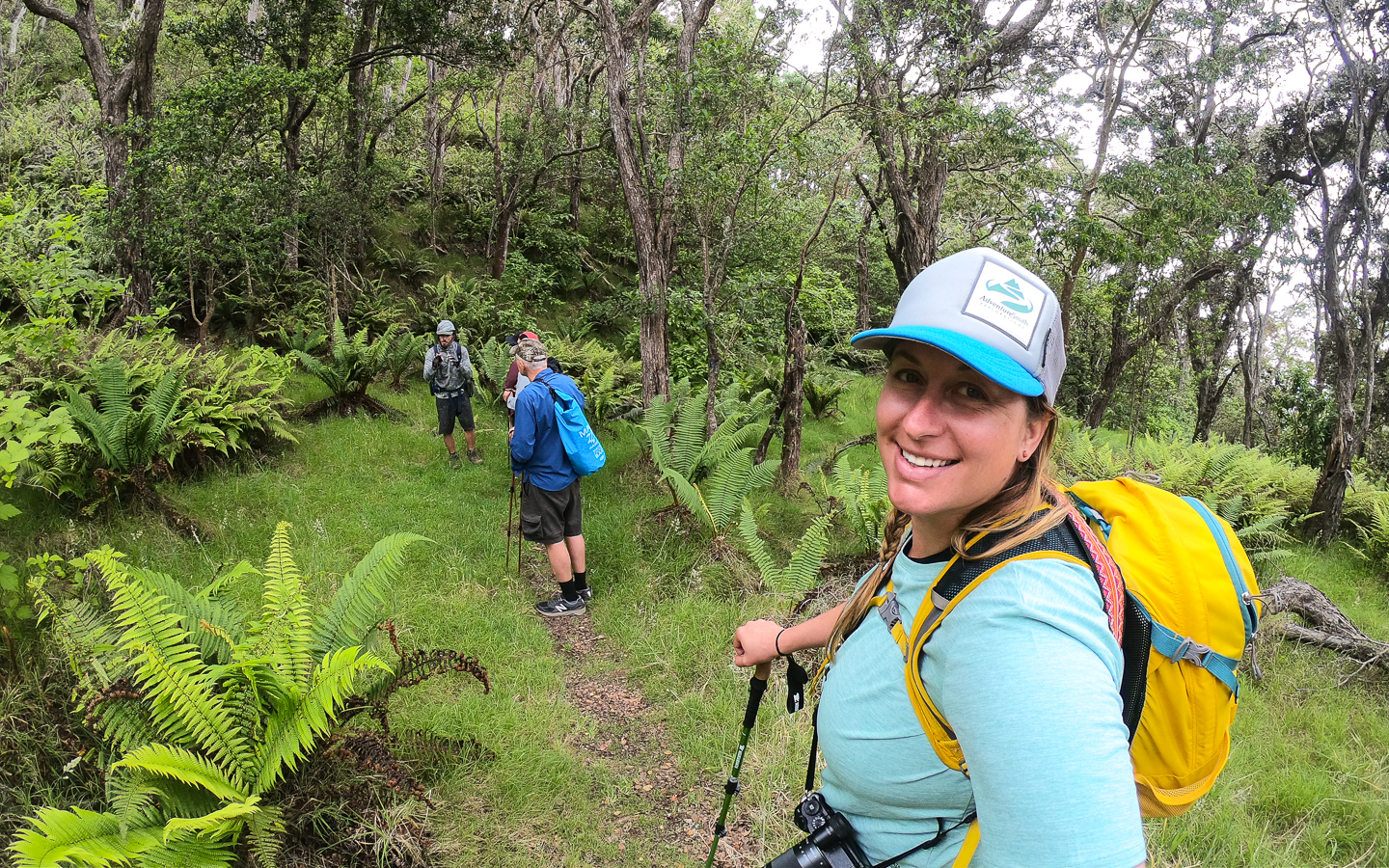 A female traveler in a blue shirt and yellow backpack looks at the camera while hiking on a tropical lush trail in the Hawaiian Islands