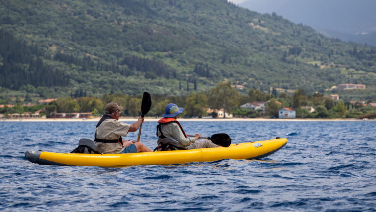 Tandem kayakers weiring beige paddle an inflatable yellow boat along lush green shores on a hot summer day in Greece.