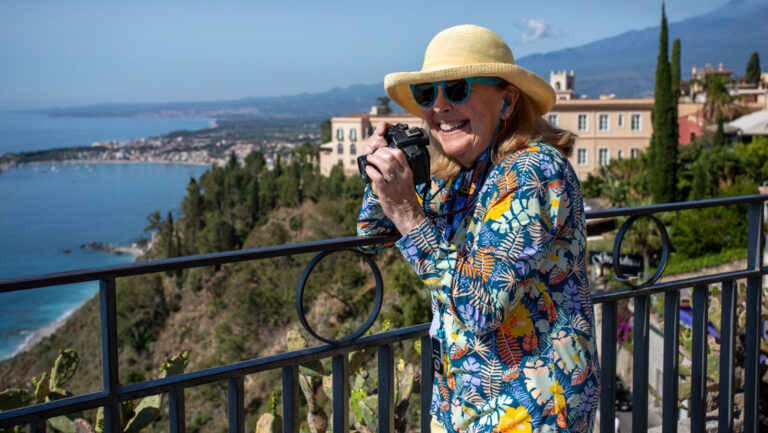 Woman in multi-color floral shirt holds camera to photograph Taormina from an overlook with a black wrought-iron fence.