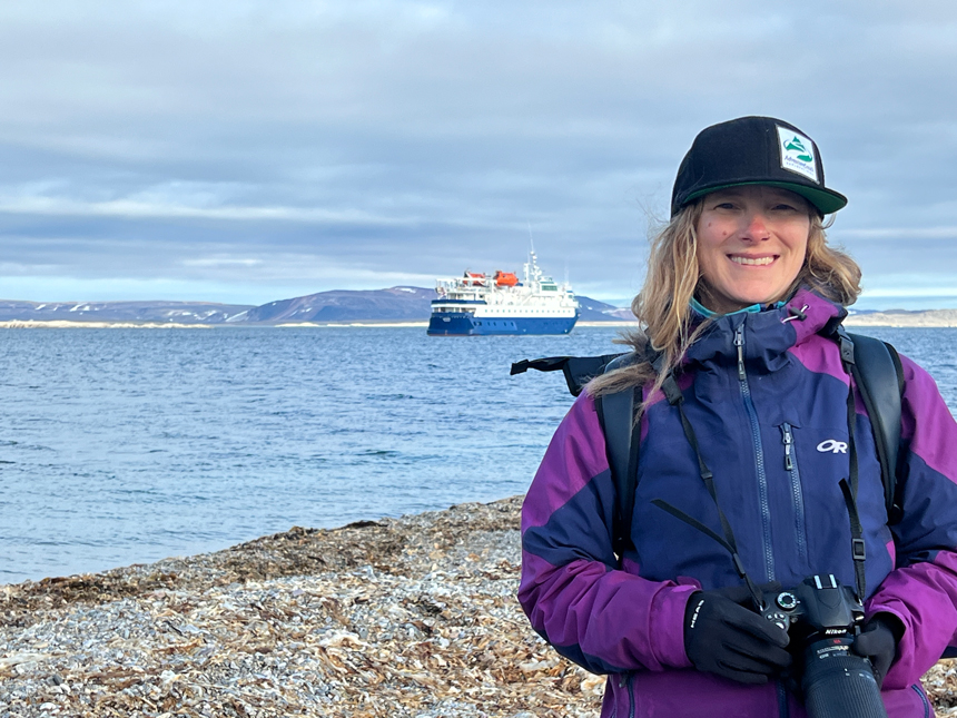 Woman in purple jacket & black baseball cap stands on shore near blue & white small ship M/S Quest in the Arctic.