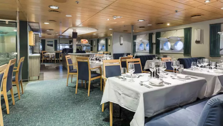 Dining room of Quest expedition ship set for dinner with white tablecloths, wood chairs, blue booths & porthole windows.