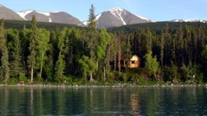 One of the cabins at the Kenai Backcountry Lodge in Alaska is just barely visible through dense forest under mountains and beyond the lake