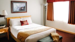 Single large bed made with white sheets and one red accent pillow in the middle next to a window at the Talkeetna Lodge in Alaska.