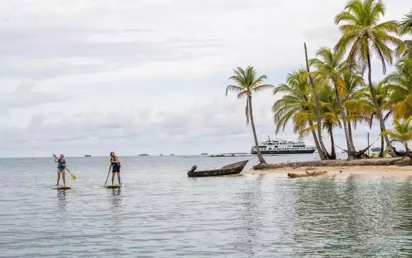 Man & woman paddle stand-up paddlebaords by shore of tiny island with white-sand beach & palm trees as ship sits nearby in Panama.