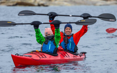 Man & girl raise kayak paddles while in a red boat during the Arctic Golden Autumn & Northern Lights trip.