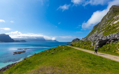 Arctic Golden Autumn & Northern Lights traveler walks a flat road by green grass & teal sea on a sunny day in Norway.