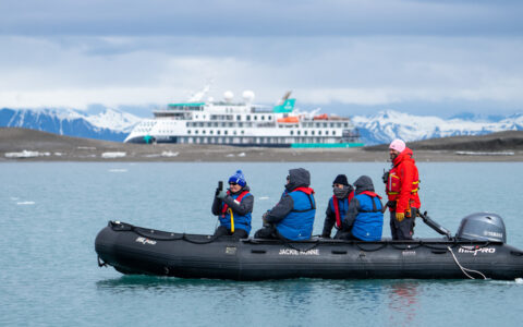 Zodiac with Arctic Golden Autumn & Northern Lights travelers moves through calm sea while guests take photos as ship sits nearby.