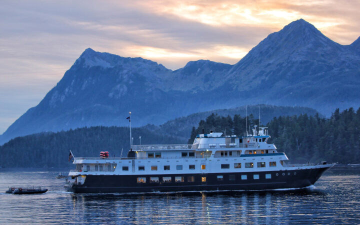 A Small cruise ship sailing through a calm bay on a beautiful night infront if stunning snowy mountains