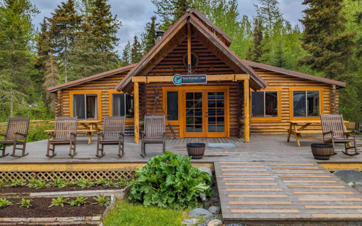 Exterior of Kenai Riverside Lodge main lodge with wood deck & rocking chairs, log cabin style, gardens & forest setting.