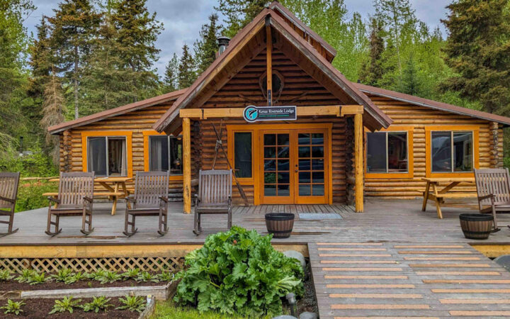Exterior of Kenai Riverside Lodge main lodge with wood deck & rocking chairs, log cabin style, gardens & forest setting.