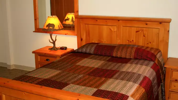 Bedroom at the King Salmon, with double wooden bedframe, checkered bedspread with brown and red color, wooden side table with small lamp and a mirror.