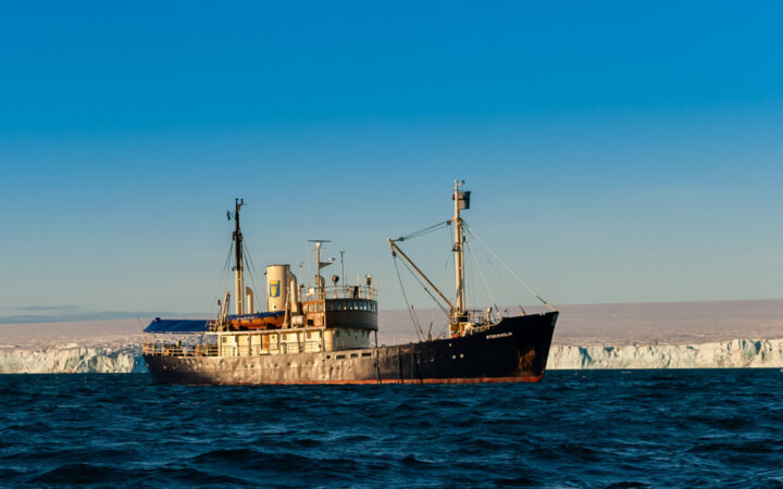 MS Stockholm ship with dark blue hull, white & wood upper decks, 2 masts & covered aft deck sits beside glacier on a sunny day.