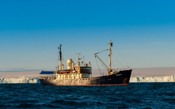 MS Stockholm ship with dark blue hull, white & wood upper decks, 2 masts & covered aft deck sits beside glacier on a sunny day.