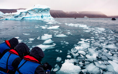 3 Northwest Passage travelers lean over side of Zodiac boat to look at iceberg bits on a cloudy day in the Canadian arctic.