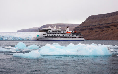 Small white expedition ship with sharp bow sits in calm water by small icebergs & tall brown mountains in the Northwest Passage.