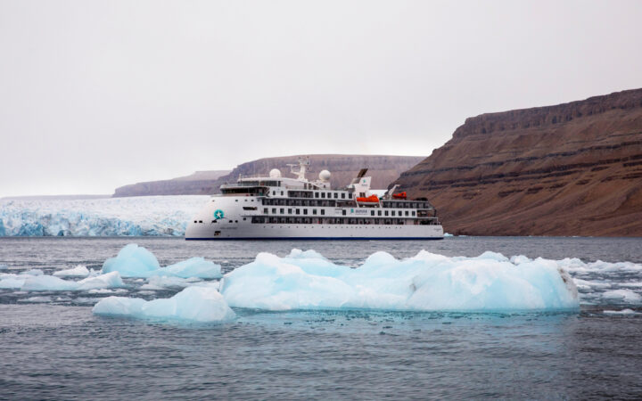 Small white expedition ship with sharp bow sits in calm water by small icebergs & tall brown mountains in the Northwest Passage.