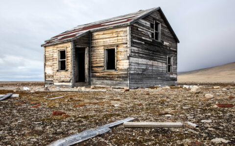 Lone weathered wood hut stands among deserted beige arctic tundra, seen on the Complete Northwest Passage Expedition.
