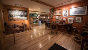 Tonglen Lake Denali lodge art gallery with framed paintings hung, crafts on wooden tables & cafe in the background.