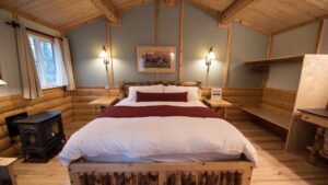 Tonglen Lake Denali lodge cabin interior with king bed, wood accents, bright windows, lamps & small cast iron stove.
