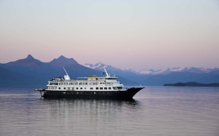 The pink and purple sunset colors the sky reflecting onto the sea, the luxury Alaska cruise ship, Safari Endeavour floats on the horizon against a purple mountain range.