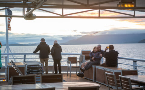 Group of travelers gather at the back of the boat to enjoy the sunset off the top deck and snap some pictures