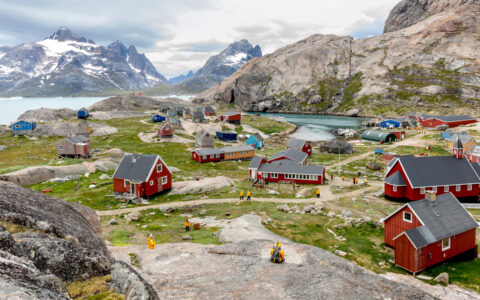 Iceland to Greenland traveler in yellow jacket sits on rock overlooking small arctic town with red buildings by the sea.