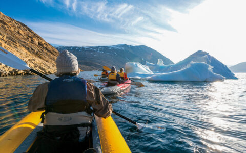 Man dressed warmly paddles front of yellow inflatable kayak beside tan rock cliffs & blue floating icebergs in Greenland.