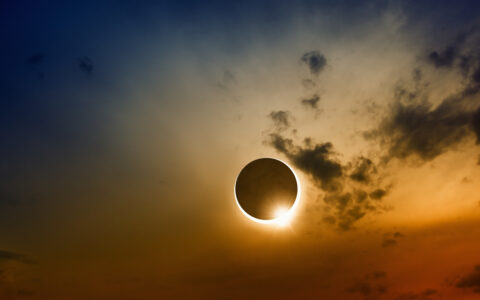 Total solar eclipse with sun nearly behind dark moon among wispy clouds & orange, red, yellow & blue sky.