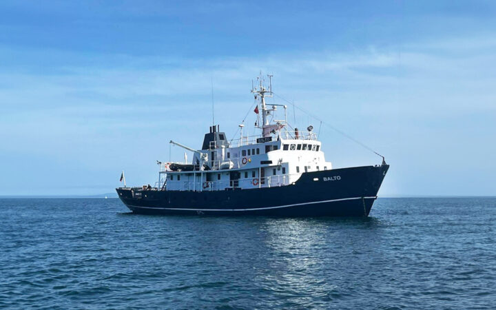 MS Balto yacht with dark blue hull & 2 white upper decks sits in calm Arctic ocean on a sunny day with blue skies.