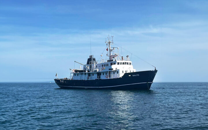 MS Balto yacht with dark blue hull & 2 white upper decks sits in calm Arctic ocean on a sunny day with blue skies.
