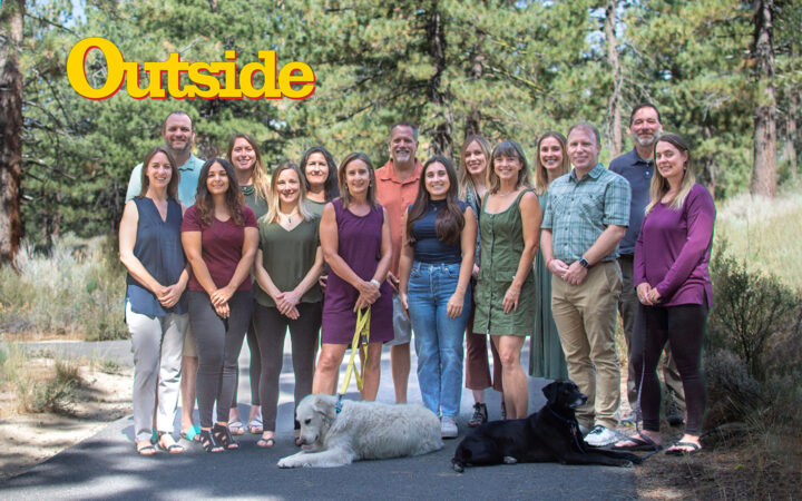 The staff of AdventureSmith Explorations. Posing outside amongst pine trees in two lines with two office dogs, one white and one black.