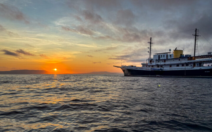 Small ship cruise pointing into the sun as it shines bright orange and yellow over the calm waters of Galapagos