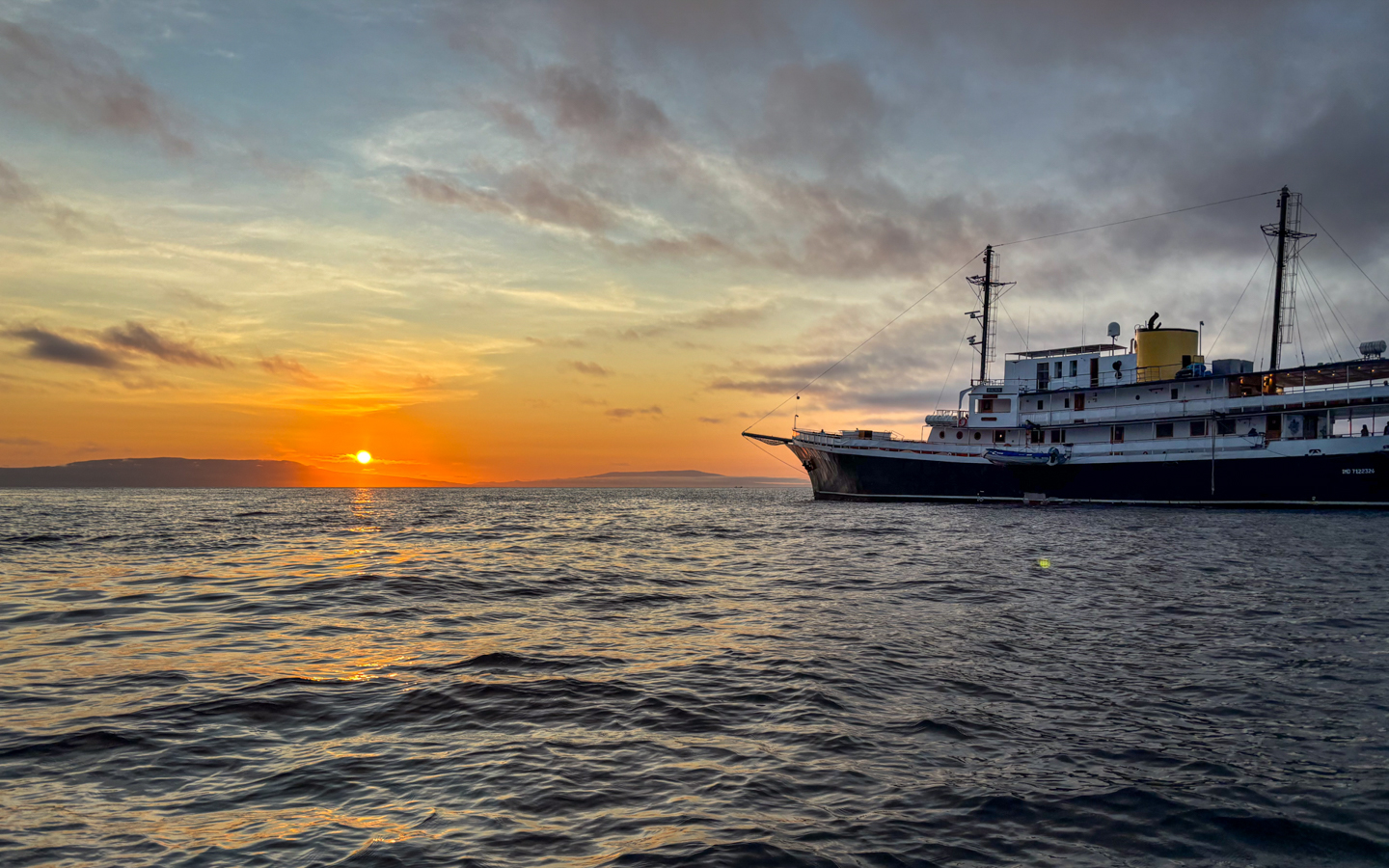 Small ship cruise pointing into the sun as it shines bright orange and yellow over the calm waters of Galapagos