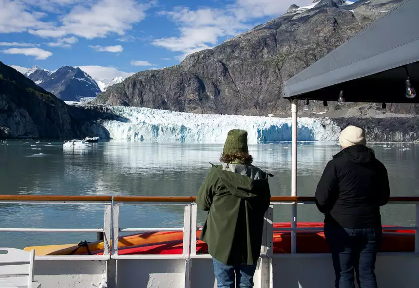 Two guests stand on bow of ship against railing and look at the glacier in front of them. 