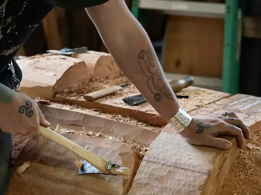 Arm with tattoo rests on wooden totem with carving tool in hand making a totem pole. 