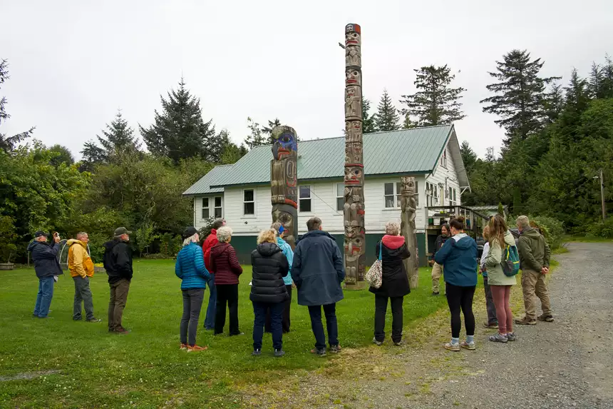 A group of Alaskan Dream Cruise guests stand in a circle around totem poles, as local village man speaks. 