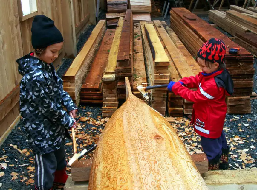 Two children wearing rain gear and beanies hold carving tools and chip away at a wooden canoe. 