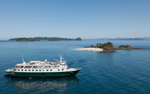 Small ship with dark green hull & white upper decks sits in calm sea offshore a tiny island on a Costa Rica adventure cruise.