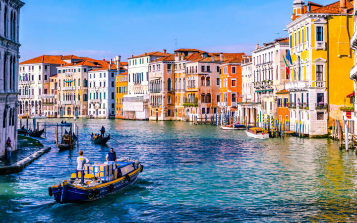 Small open-air boat motors through turquoise sea among tall, colorful buildings with terra cotta roofs in Venice, Italy.