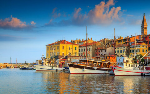 Small wooden boats with white hulls sit in calm harbor by colorful buildings & belltower in Rovinj, Croatia.