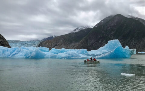 Small tender boat with Alaskan Story charter cruise guests slowly motors past massive blue iceberg chunks on a cloudy day.
