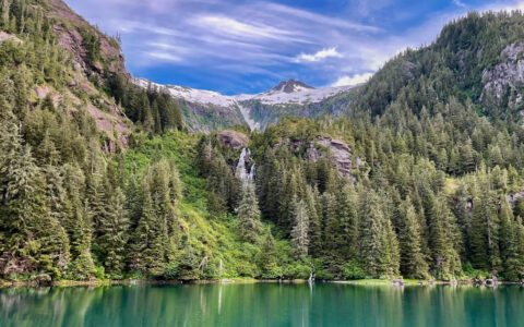 Scenic southeast Alaska with verdant mountains with granite peeking out & a waterfall spilling into turquoise sea.