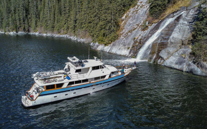 Aerial view of Alaskan Story yacht with blue & white exterior sitting in calm water in front of water cascading over rock.