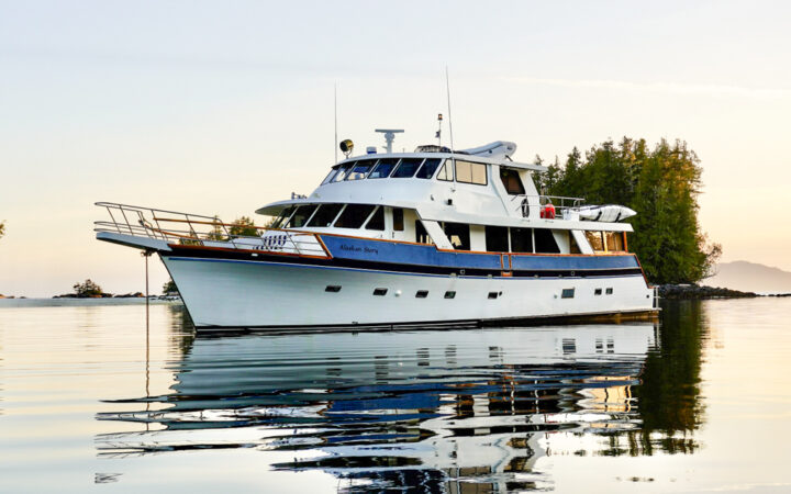Alaskan Story yacht with 3 decks, kayaks, paddleboards & blue & white exterior paint sits in glassy water at dusk.