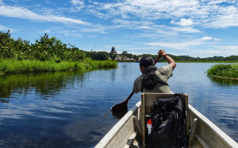 guide canoeing on a lake with blue sky, green rainforest and eco lodge in the background on an Amazon trip.