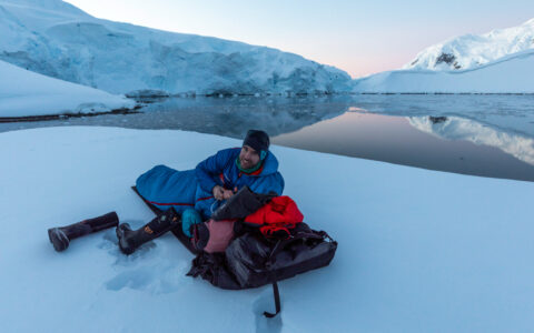 Man lays in down sleeping bag on untouched snowfield by glassy water & snowy mountains at dusk on the Antarctica Active cruise.