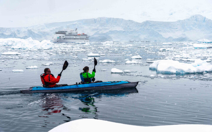 Tandem kayakers in red & green jackets paddle an inflatable blue kayak among icy waters by white expedition ship.