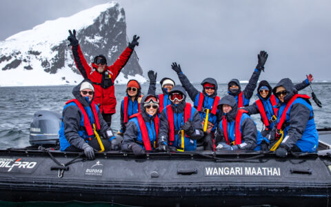 Black Zodiac boat with Antarctica Active cruise guests in blue jackets & red PFDs waving near a cliff on a cloudy day.