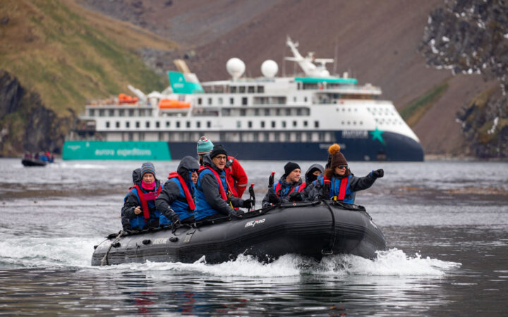 Zodiac boat with Antarctica Complete cruise guests moves through calm water with small ship in teal, blue & white behind.