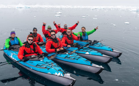 4 tandem inflatable teal kayaks sit side-by-side in calm sea as their Antarctica Complete cruise guests smile & wave.